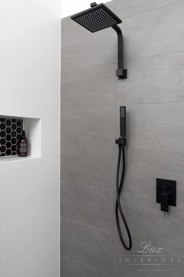 Modern bathroom shower installation featuring a matte black square rainfall showerhead, handheld shower, and black controls mounted on large light-grey wall tiles. A recessed niche with black hexagonal tile backing provides built-in storage for toiletries.