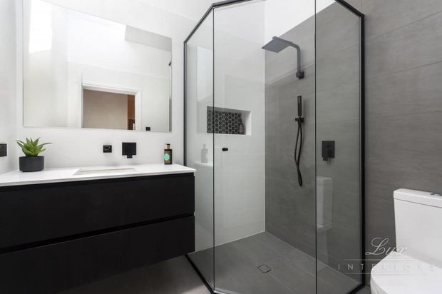 Contemporary monochrome bathroom featuring a floating matte-black vanity with white countertop, large frameless mirror, black fixtures, and a spacious walk-in shower enclosed by clear glass with dark framing. Grey wall and floor tiles create a clean, refined finish.