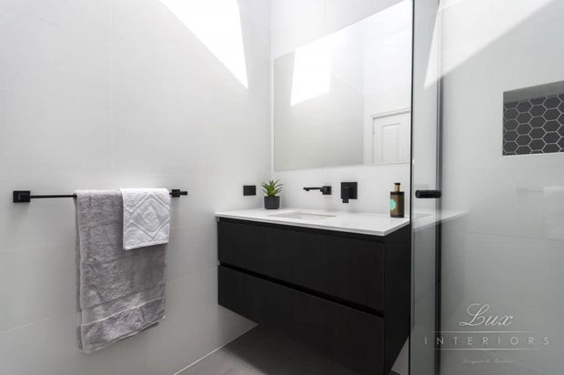 Bright contemporary bathroom featuring a wall-mounted matte black vanity with a white benchtop, large mirror, black tapware and fixtures, white tiled walls, a black-framed glass shower enclosure, and a small dark hexagonal-tile shower niche.