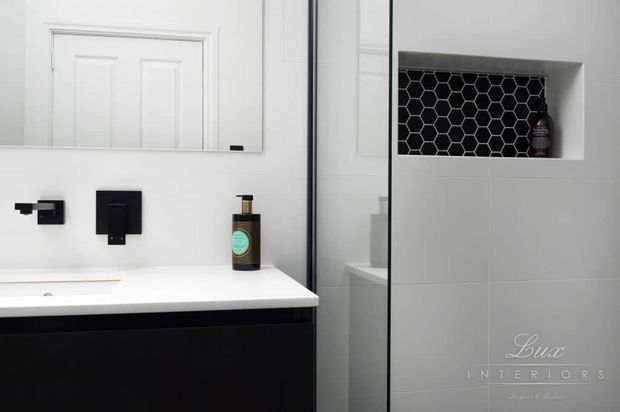 A compact contemporary bathroom vanity with a white integrated basin and benchtop, black wall-mounted tapware and cabinetry, a large mirrored cabinet, and a recessed wall niche lined with black hexagonal tiles. The space uses a restrained monochrome palette with subtle decorative detailing.