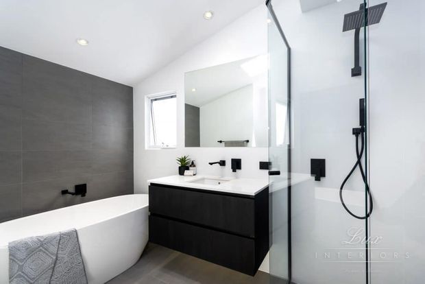 Contemporary bathroom with a freestanding white bathtub, floating dark vanity, white benchtop, wall-mounted black tapware, large mirror, and frameless glass shower enclosure. Grey tiled walls and floor create a clean, monochromatic scheme with recessed ceiling lighting.
