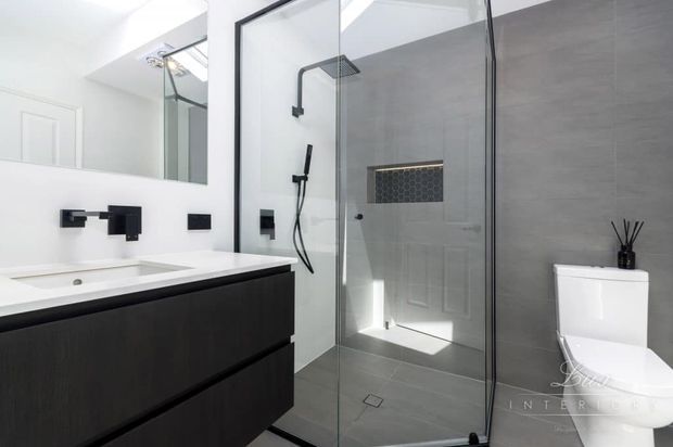 Contemporary bathroom featuring a floating dark vanity with a white integrated basin, black wall-mounted tapware, large frameless glass shower enclosure, grey tiled walls and floor, a recessed hexagonal tile niche, and a white toilet. The space is bright, crisp, and minimalist.