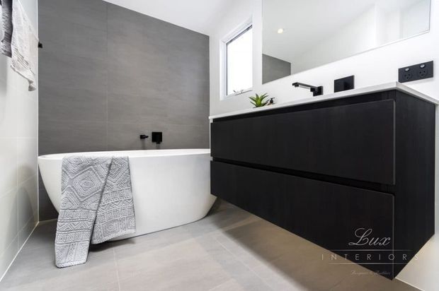 Contemporary bathroom with a freestanding white bathtub against a large grey tiled feature wall, paired with a floating matte-black vanity, black tapware, and a white countertop. A wall mirror, small window, recessed lighting, and grey floor tiles create a clean, high-contrast modern fit-out.