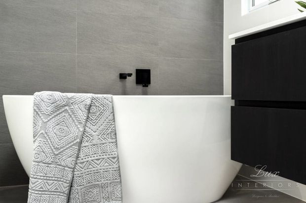 A contemporary bathroom featuring a freestanding white bathtub against large grey stone-look wall tiles. Matte black wall-mounted bath fixtures and a dark floating vanity provide contrast, while a patterned grey-and-white towel hangs over the tub.