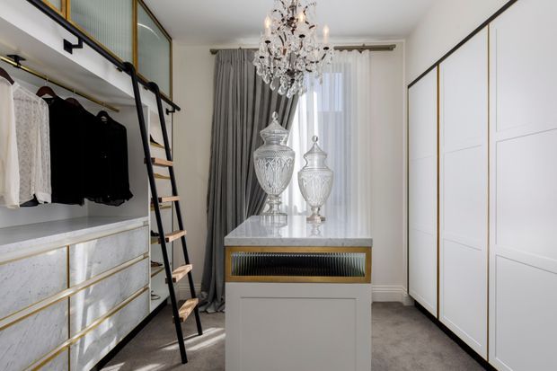 A refined walk-in wardrobe with full-height white built-in cabinetry, open hanging rails, marble-look drawer fronts with brass trim, and a central island topped in white stone. A black metal ladder provides access to upper storage. The room has grey carpet, floor-to-ceiling sheer and charcoal curtains, and a decorative crystal chandelier, creating a luxurious, tailored dressing space.