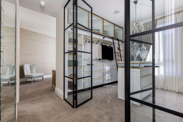 A refined walk-in wardrobe with floor-to-ceiling black-framed glazed steel-style partition doors, extensive light cabinetry, hanging rails and open shelving. The wardrobe opens toward a carpeted bedroom or dressing area, visible directly and in reflections, with a chandelier and sheer curtains adding an elegant finish.