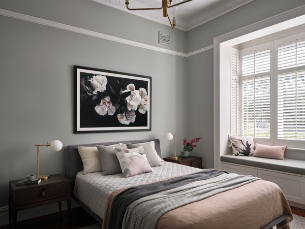 A refined bedroom with a dark upholstered bed, layered neutral bedding, blush and grey cushions, and a folded textured throw. The room has muted grey walls, white decorative cornice and trim, a floral artwork above the bed, contemporary bedside tables and globe lamps, plus a bright bay window with plantation shutters and a built-in window seat. The overall look is calm, elegant, and softly traditional with contemporary furnishings.