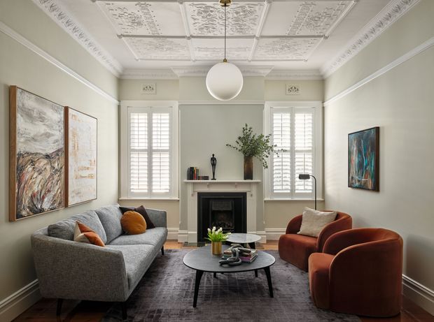 A refined living room with a grey sofa, rust-toned armchairs, layered round coffee tables, a central fireplace with mantel, plantation shutters, framed artwork, and a highly detailed pressed-tin decorative ceiling. The palette is soft grey, off-white, charcoal, and warm rust accents, creating a polished yet inviting atmosphere.