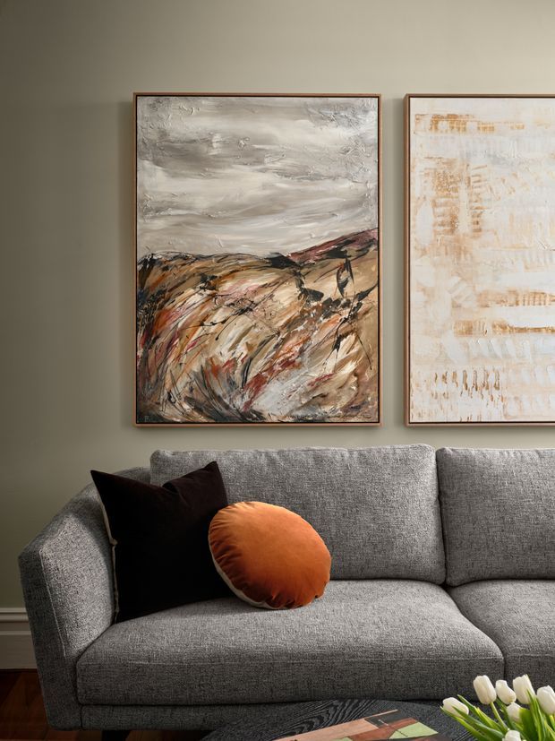 Close-up interior view of a contemporary living room featuring a grey upholstered sectional sofa with black and rust-orange cushions. Two large abstract landscape paintings in slim timber frames hang on a muted olive-grey wall above the sofa. Dark timber flooring and a small portion of a coffee table are visible, with flowers in the foreground. The styling is warm, refined, and art-focused.