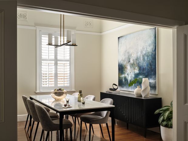 A refined dining room viewed through an open doorway, featuring a rectangular dining table with upholstered chairs, a large abstract artwork, a black ribbed sideboard, a contemporary linear pendant light, plantation shutters, warm timber flooring, and neutral cream walls. Decorative vases and indoor greenery add restrained accents.