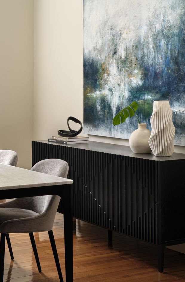 A refined dining area featuring a dark, fluted sideboard against a warm neutral wall. A large abstract blue, white, and grey artwork hangs above the sideboard, which is styled with sculptural vases, a small leafy plant, books, and a black abstract ornament. In the foreground, a light stone or pale tabletop with upholstered grey dining chairs sits on a warm timber floor. The interior has a sophisticated, moody, contemporary feel with strong vertical ribbed detailing.