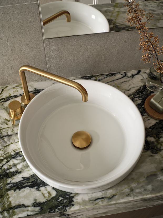 Close-up view of a refined bathroom vanity featuring a white round vessel basin, brushed brass curved wall-mounted faucet and matching drain, set into a dramatic veined marble countertop. A large mirror reflects the basin and faucet, while a small decorative branch and soap accessories sit to the side.