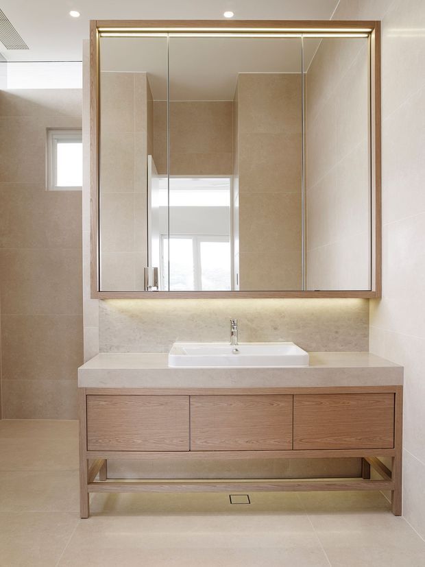 Bright contemporary bathroom featuring a wall-mounted or raised timber vanity with three drawers, a white rectangular basin, pale stone benchtop and matching tiled walls and floor. A large mirrored cabinet with a timber frame spans the vanity, with concealed warm LED lighting beneath it. The room has clean lines, recessed ceiling lights, and a small high window.