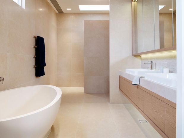 Bright contemporary bathroom featuring a freestanding white bathtub, large-format beige wall and floor tiles, a floating light-wood vanity with white benchtop and integrated basins, mirrored cabinetry, a wall-mounted towel rail, and a skylight. The room has a clean, spacious and refined appearance.