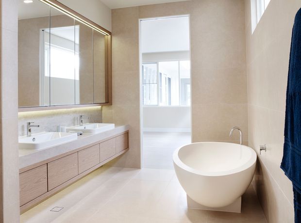 Bright contemporary bathroom featuring a freestanding oval bathtub, wall-mounted double vanity with pale timber cabinetry, large illuminated mirrored cabinet, pale stone-look wall and floor finishes, and a glass-paneled doorway leading to a bright adjoining space.