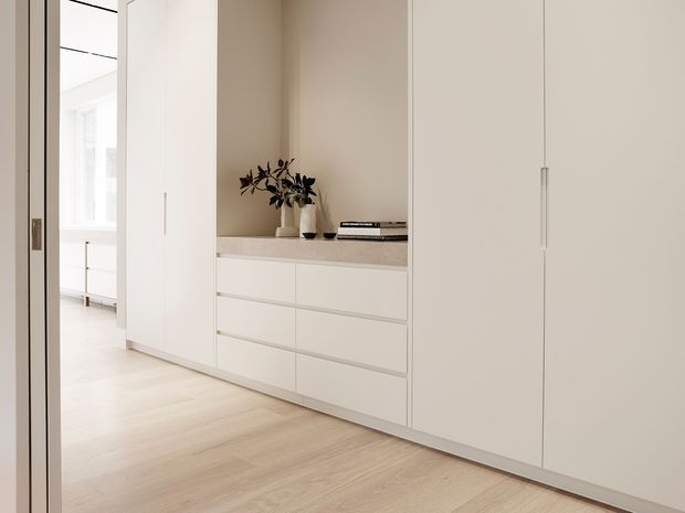 A bright, minimalist built-in storage wall viewed from a hallway or adjacent room. The pale cabinetry features tall flush doors, integrated slim handles, lower drawers, and an open display niche with a light benchtop holding a vase and books. Light timber flooring and soft off-white finishes create a calm, understated interior.