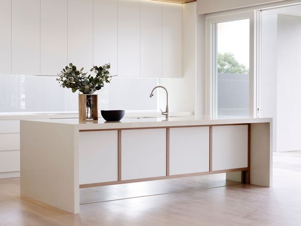 A bright, minimalist kitchen featuring a large white island with integrated cabinetry and a slim timber-toned trim. The island includes a pale benchtop and curved metal tap, while full-height white cabinetry lines the rear wall. Light timber flooring, concealed warm LED lighting, and large glazed sliding doors create an airy connection to the exterior.