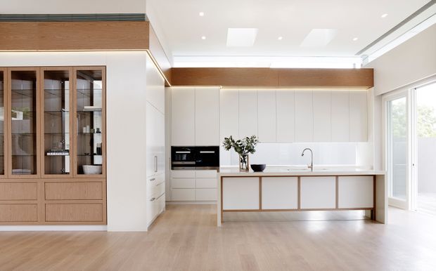 Bright contemporary kitchen with a large white island, integrated white cabinetry, pale timber flooring, and extensive timber-faced built-in joinery. The space includes a glazed display cabinet, concealed appliances, recessed ceiling lighting, a timber ceiling bulkhead, and large glazed doors opening toward the exterior.