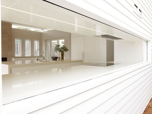 Bright, minimalist kitchen viewed from outside through a large open service window. The space features glossy white built-in cabinetry, a broad white island or benchtop, integrated appliances, a stainless or chrome gooseneck tap, pale stone or tiled walls, plantation shutters, and a white horizontal weatherboard exterior around the opening. A small indoor plant adds the only notable visual contrast.