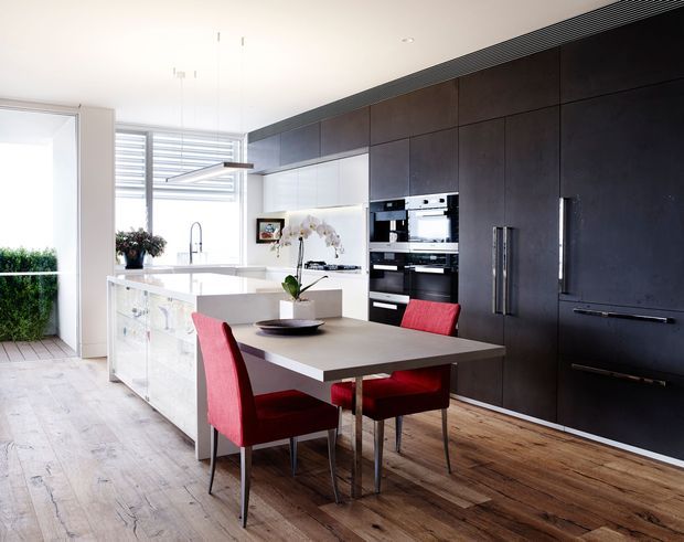 A bright, contemporary open-plan kitchen and dining area. The kitchen features a large white island with an integrated sink, dark full-height cabinetry with integrated stainless-steel ovens and appliances, pale timber flooring, and a compact dining table with vivid red upholstered chairs. Large glazing and louvred window screening bring in natural light, while pendant lighting hangs above the island.