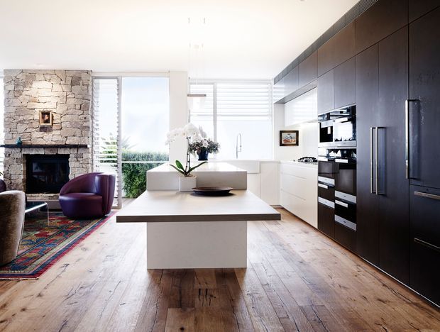 Bright open-plan kitchen and living area with a large central island, white cabinetry, dark full-height kitchen joinery, integrated appliances, timber flooring, and a substantial natural stone fireplace. Large windows with horizontal blinds bring in abundant daylight, while a purple lounge chair and patterned rug add colour.