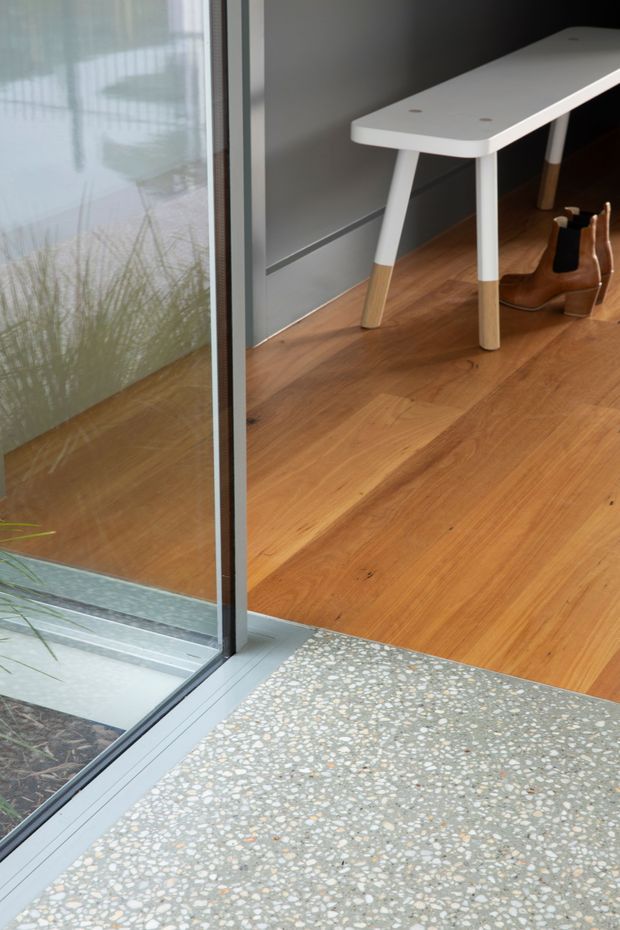 Close-up architectural interior detail showing a threshold between a terrazzo-finished entry or exterior slab and a warm timber-floored room. A large glazed sliding door with slim dark metal framing separates the spaces. Inside, a simple white-top timber-legged table and wooden chair are partially visible against a dark grey wall.