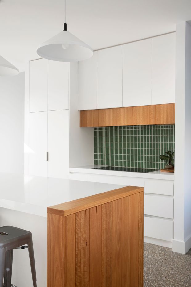 Bright minimalist kitchen with full-height white cabinetry, a warm timber island and matching timber overhead feature cabinetry, green vertical tile splashback, white benchtops, pendant lighting, and a terrazzo-like floor.
