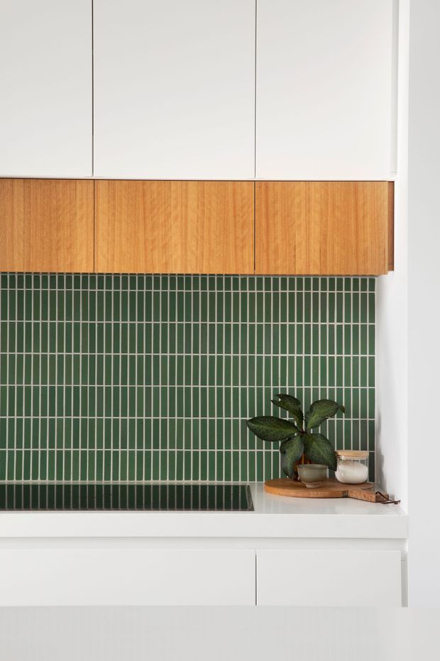 Close-up view of a compact contemporary kitchen with white overhead and lower cabinetry, warm timber-grain cabinet fronts, and a dark green vertically arranged tiled splashback. A black cooktop is visible below, with a small timber tray and potted plant on the benchtop.
