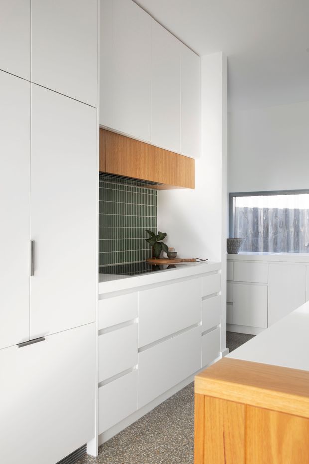 A bright, compact contemporary kitchen with extensive matte white full-height cabinetry, integrated drawers, a warm timber overhead cabinet and kitchen island, and a dark green vertically ribbed tile splashback. A small timber shelf with simple decor sits above the cabinetry, while a window brings in natural light. The floor appears to be terrazzo or pebble-flecked concrete.