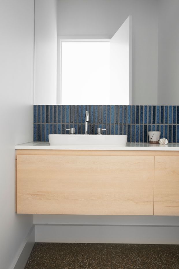 A bright, minimalist bathroom vanity with a wall-mounted pale timber cabinet, white integrated countertop and vessel basin, chrome tapware, and a striking blue vertically patterned tiled splashback. A frameless mirror or bright window-like opening sits above the vanity, and the floor appears to be dark terrazzo.
