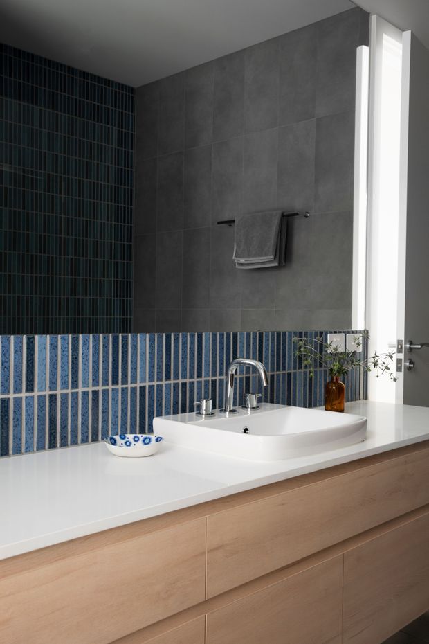 A contemporary bathroom vanity with a white countertop and integrated ceramic basin, brushed metal tapware, light timber drawer cabinetry, and a tiled backsplash. The lower backsplash uses vertically oriented blue patterned tiles, while the upper wall is finished in large charcoal-grey tiles. A narrow vertical window brings in strong natural light beside a towel rail.