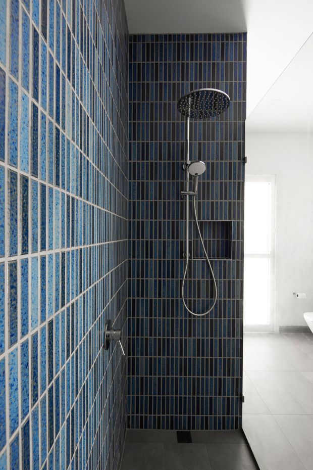 A contemporary bathroom shower enclosure lined with narrow, vertically oriented blue and dark grey tiles. The shower features a large round rainfall showerhead, handheld shower, recessed niche, and dark grey tiled floor. A bright white bathroom with a window and partial freestanding basin is visible beyond the enclosure.