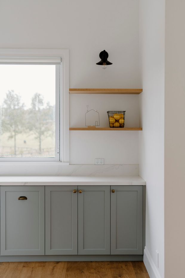 A bright, compact kitchen corner with muted grey-green shaker cabinetry, a white marble-look benchtop and splashback, warm timber flooring, and open timber shelves. A black wall sconce, wire fruit basket, and small line-art house add restrained decorative details beside a large window.