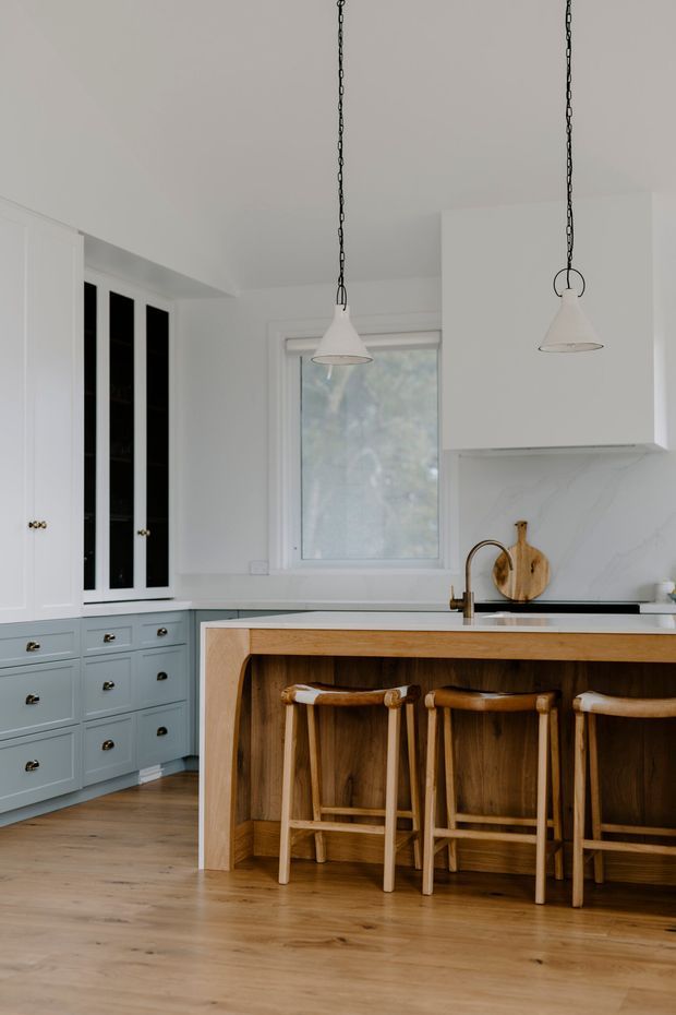 A bright, refined kitchen with pale blue-grey shaker cabinetry, white walls and ceiling, timber flooring, and a large timber island with a light stone benchtop. Three timber bar stools sit beneath the island overhang, while two white pendant lights hang from dark chains. Black-framed glazed cabinet doors, brass-toned hardware, a white marble-look splashback, and a window overlooking greenery complete the space.