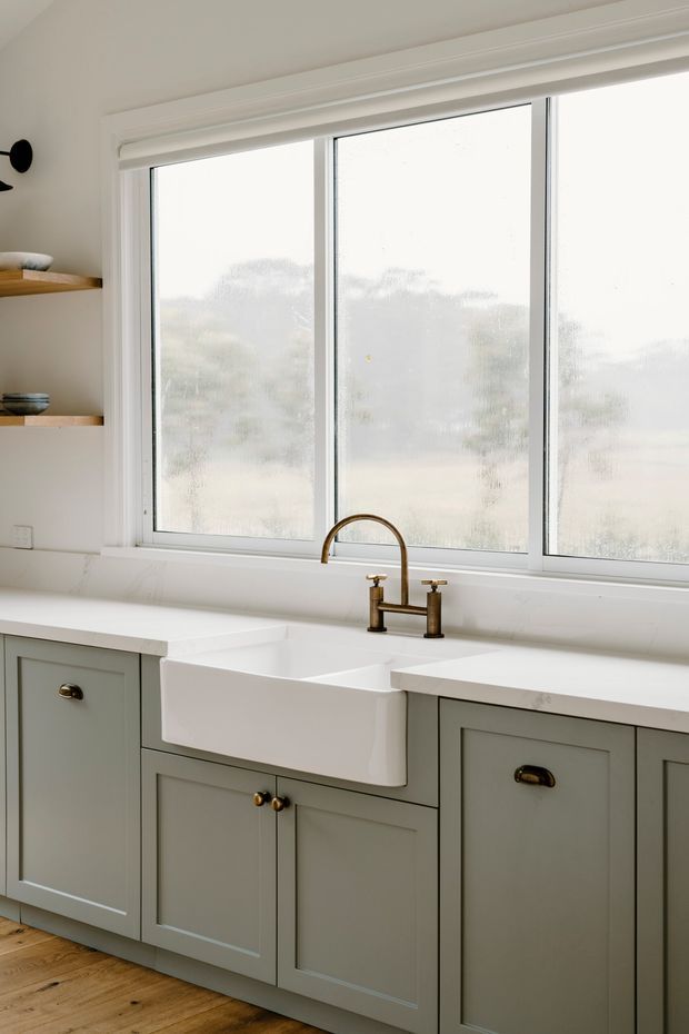 Bright, calm kitchen vignette featuring a large three-panel window, white farmhouse apron-front sink, brushed brass bridge faucet, pale sage-grey shaker cabinetry with brass hardware, white benchtops and splashback, open timber display shelves, and timber flooring.