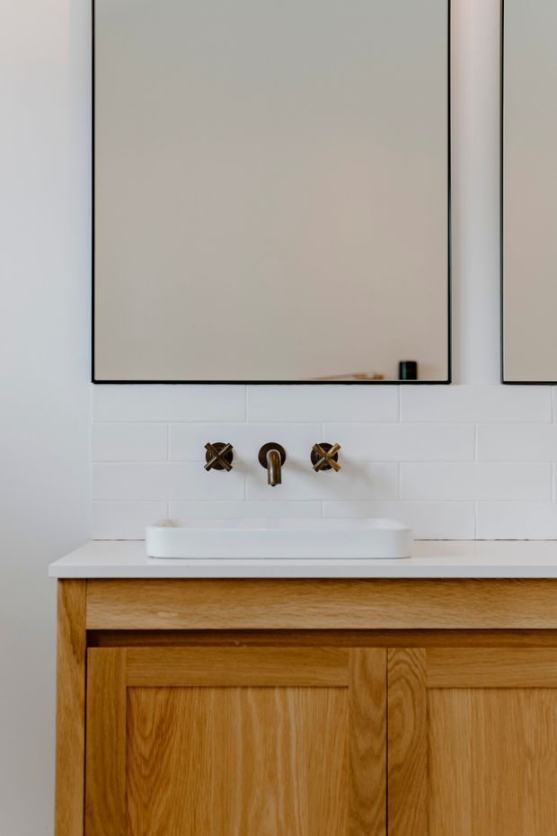 Close-up view of a refined bathroom vanity featuring a white vessel basin, wall-mounted bronze tapware, white subway-tile splashback, pale stone or solid-surface benchtop, and warm oak cabinetry with framed panel doors. A large frameless or dark-edged mirror is mounted above the vanity.