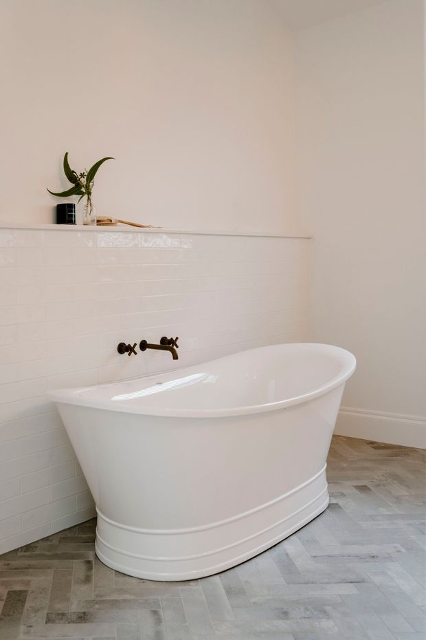 A bright, refined bathroom featuring a freestanding white bathtub positioned against a white glazed subway-tile half wall. A dark bronze wall-mounted bath filler provides contrast, while pale grey stone-look herringbone flooring and a simple ledge with a small plant complete the calm, minimalist interior.