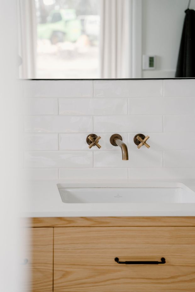 Close-up view of a refined bathroom vanity featuring a white rectangular inset basin, wall-mounted aged brass cross-handle tapware, glossy white subway-tile splashback, and a light timber vanity with black pull handle. A large mirror and window are visible above.