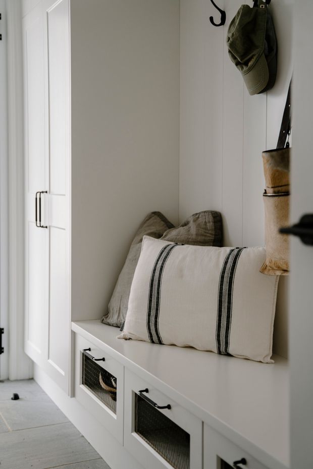 A compact, calm entryway or mudroom with built-in white cabinetry, a cushioned window or bench seat, paneled walls, open wire-front storage drawers, and black hooks holding hats and bags. Neutral linen pillows and light grey flooring create a soft, understated farmhouse-inspired look.