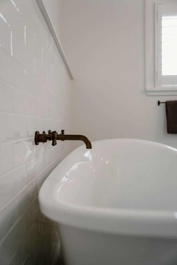 Close-up photograph of a white freestanding bathtub in a bright bathroom, set against glossy white tiled walls. A wall-mounted aged bronze or dark metal faucet projects over the tub, with a small louvered window and dark towel visible in the background. The scene has a clean, understated traditional feel.