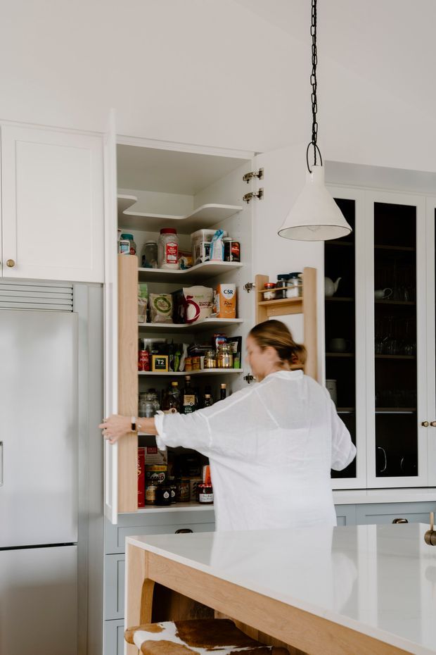 A bright, refined kitchen with white shaker-style cabinetry, a concealed walk-in pantry cupboard filled with provisions, glass-front upper cabinets, and a pale stone island with a light timber base. A large white pendant hangs above the island, while a person opens the pantry door. Stainless-steel refrigeration and warm timber details add contrast.