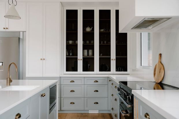 Bright, refined white kitchen with shaker-style cabinetry, glazed display cupboards, brass hardware, pale stone benchtops, a large central island, freestanding stainless-steel range cooker, and a window above the cooking area. The space has a clean, classic fitted design with warm timber flooring and dark accents inside the display cabinetry.