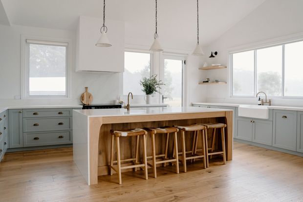 Bright, spacious kitchen with a large central island featuring integrated timber seating and a light benchtop. Pale blue-grey shaker cabinetry, white walls, timber flooring, open shelving, pendant lights, and generous windows create a calm, airy interior.