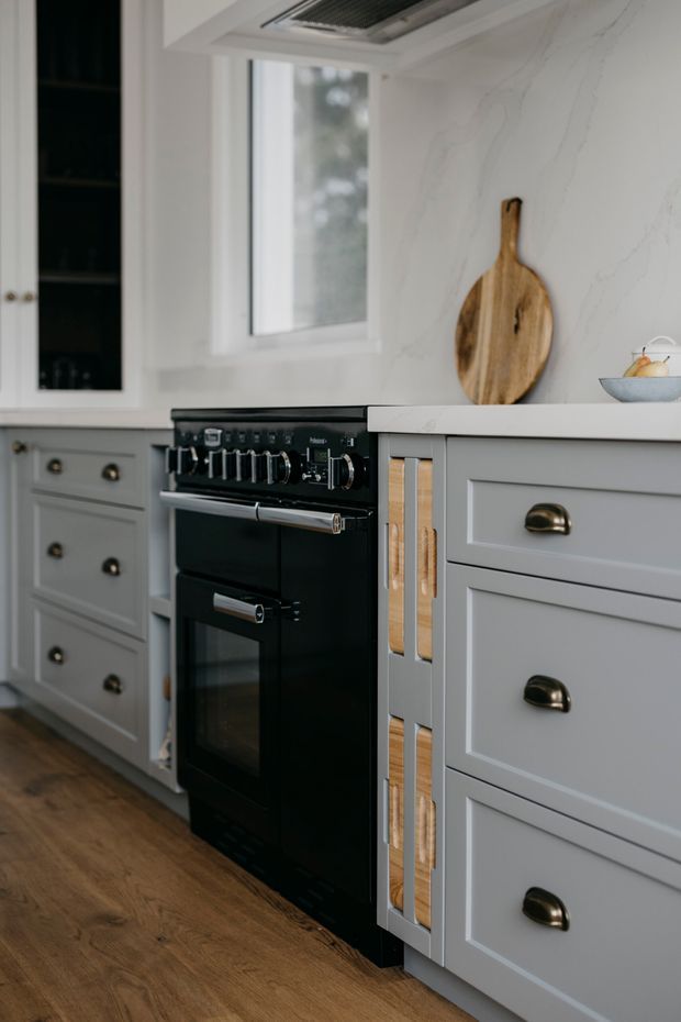 A close-up view of a refined kitchen featuring grey shaker-style cabinetry with dark cup handles, a black freestanding range cooker, white marble-look benchtops and splashback, timber flooring, and a window above the counter. A timber cutting board and small bowl sit on the benchtop, while open shelving and a rangehood are visible in the background.