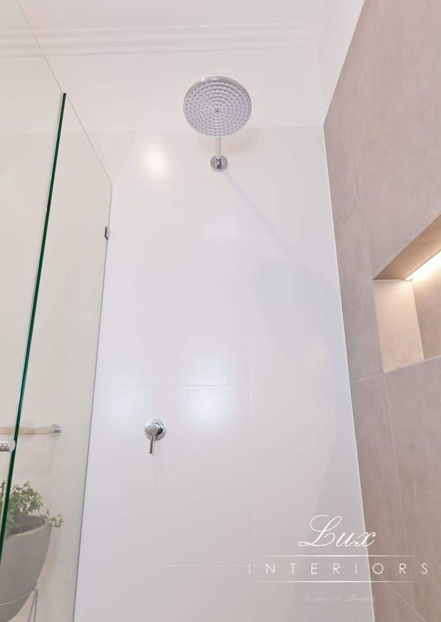 Close-up view of a contemporary bathroom shower area with large glossy white wall tiles, a round chrome rain showerhead, concealed shower controls, and a frameless glass shower screen. Neutral grey-beige tiled surfaces and recessed lighting are visible at the right edge.