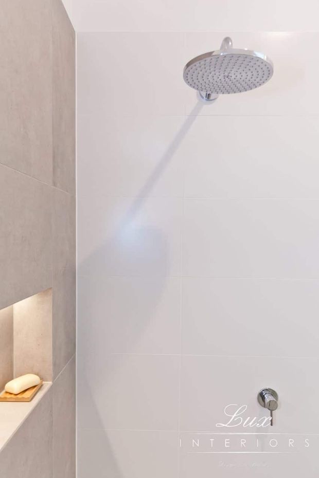 Close-up view of a contemporary shower enclosure featuring a large round chrome rain showerhead, white wall tiles, and a contrasting beige-grey tiled wall with a recessed shelf holding soap. The space is bright, clean, and minimal.