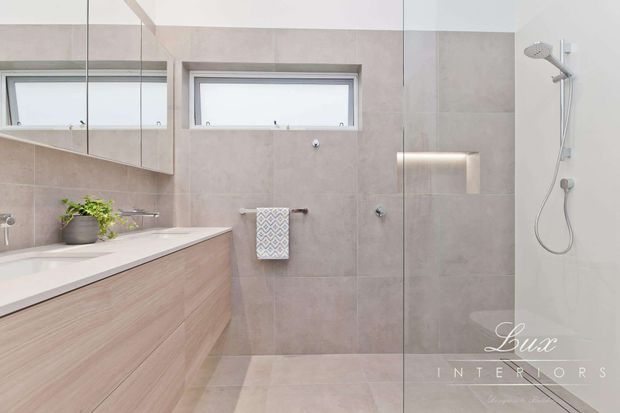 Bright contemporary bathroom featuring a frameless glass shower enclosure, wall-mounted shower, large-format beige wall and floor tiles, pale timber-look vanity cabinetry, white benchtop, mirrored wall cabinetry, recessed shelving, and a small high window. The space has a clean, restrained and softly neutral aesthetic.