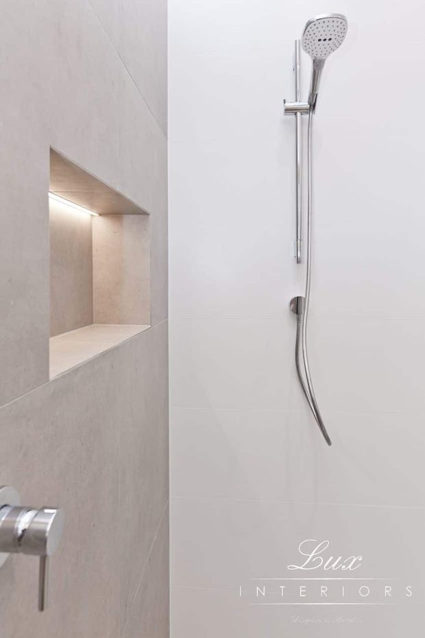 Minimal contemporary shower area with a chrome handheld shower rail and controls mounted against large white wall tiles. A recessed niche with integrated lighting is finished in pale beige stone-look tile, creating a clean, refined bathroom interior.