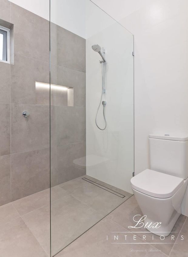 Modern minimalist bathroom featuring a frameless glass walk-in shower, large light-grey tiled walls and floor, recessed illuminated niche, chrome shower fittings, and a white wall-adjacent toilet. The space is bright, clean, and neutral with streamlined contemporary detailing.