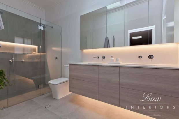 Contemporary bathroom with a walk-in glass shower, wall-hung toilet, large floating double vanity, mirrored wall cabinetry, and integrated warm LED lighting. The space uses pale grey tiled surfaces, light wood-look vanity cabinetry, and minimalist black and chrome fixtures.